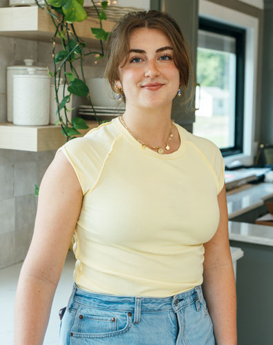 A young woman smiling in a kitchen. She is wearing a yellow top and jeans.