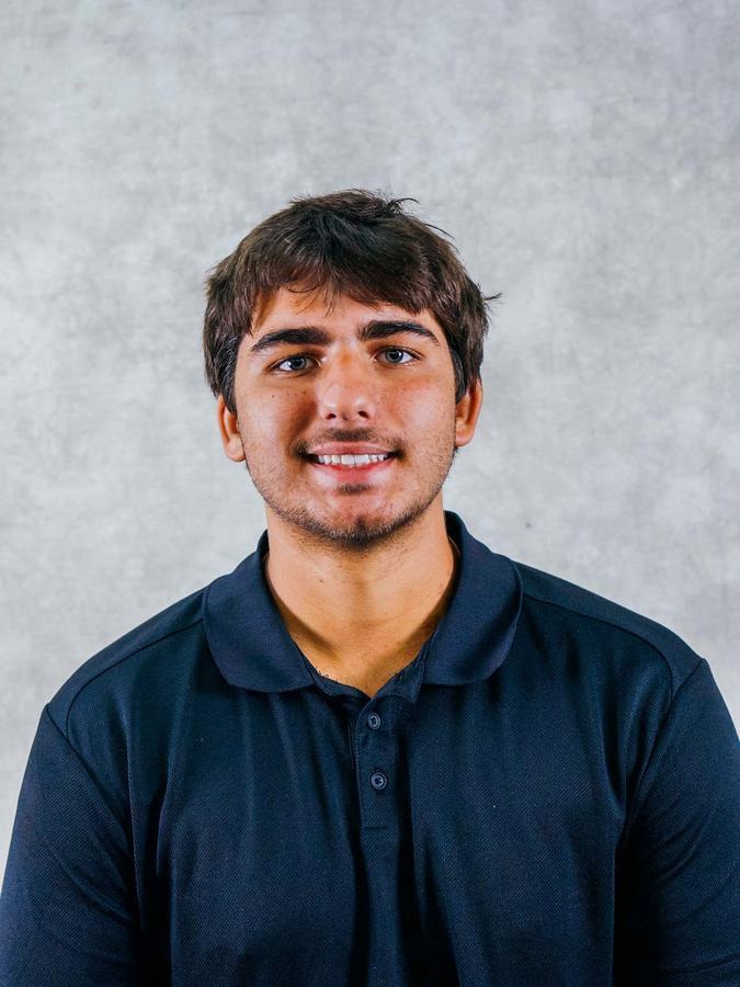 A smiling young man with short dark hair and a light beard posing in a navy blue polo shirt against a plain gray background.
