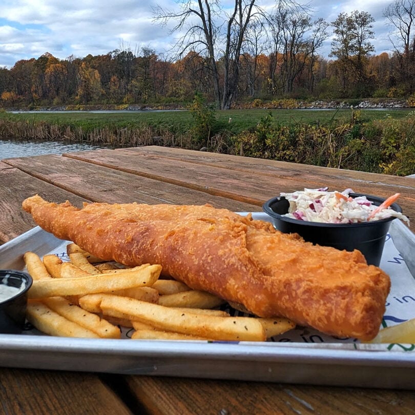 A plate of fish and chips with coleslaw and tartar sauce on a wooden table overlooking a body of water. The food looks freshly prepared and appetizing.